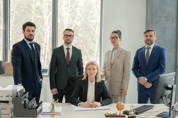 Serious mature female chef of law firm surrounded by lawyers sitting with documents at desk in modern office