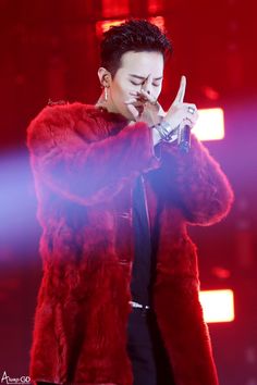 This contains an image of G-Dragon's  wearing a red fur coat and holding his  hand up to her mouth