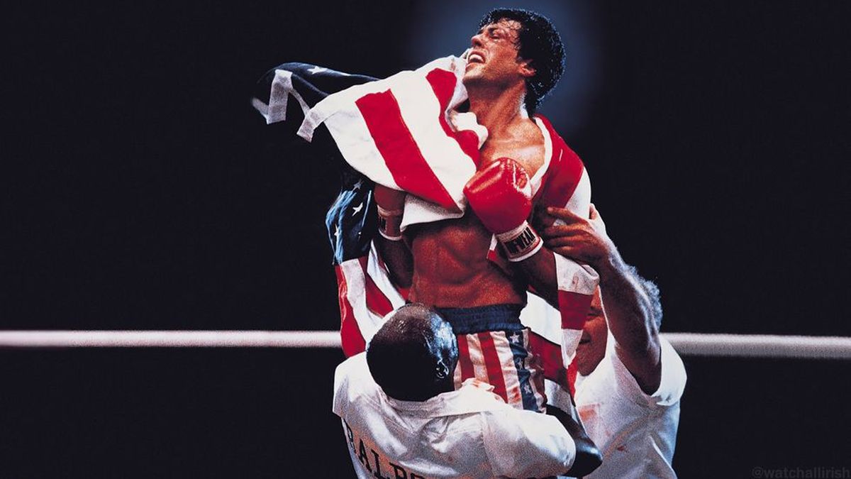 Sylvester Stallone en su papel de Rocky Balboa, envuelto en una bandera estadounidense, levantado en el ring por sus entrenadores tras una pelea. La emoción y el esfuerzo son visibles en su rostro, con guantes de boxeo rojos y pantalones de rayas patrióticas.