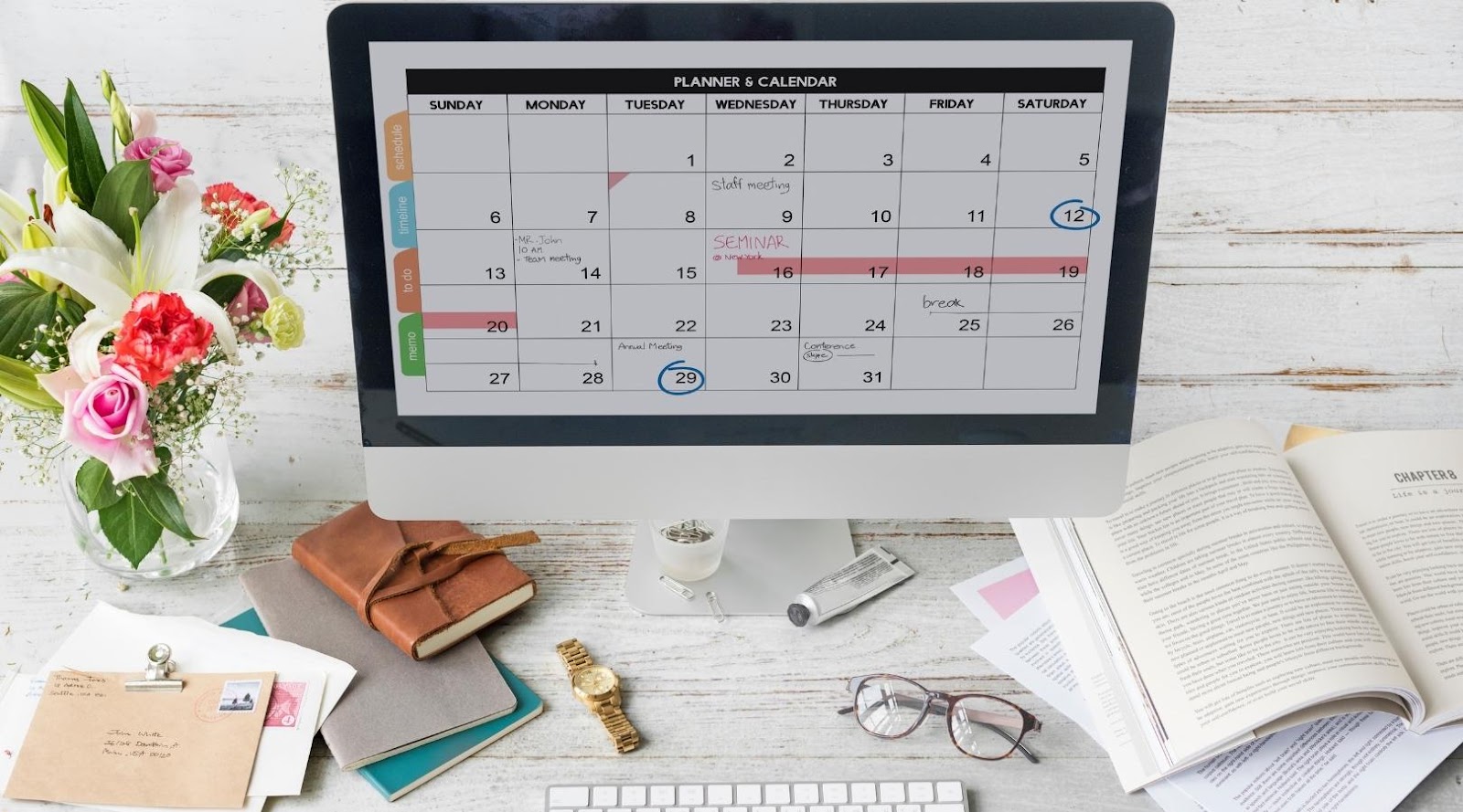 Computer displaying a digital calendar on a desk with flowers, notebooks, and an open book.