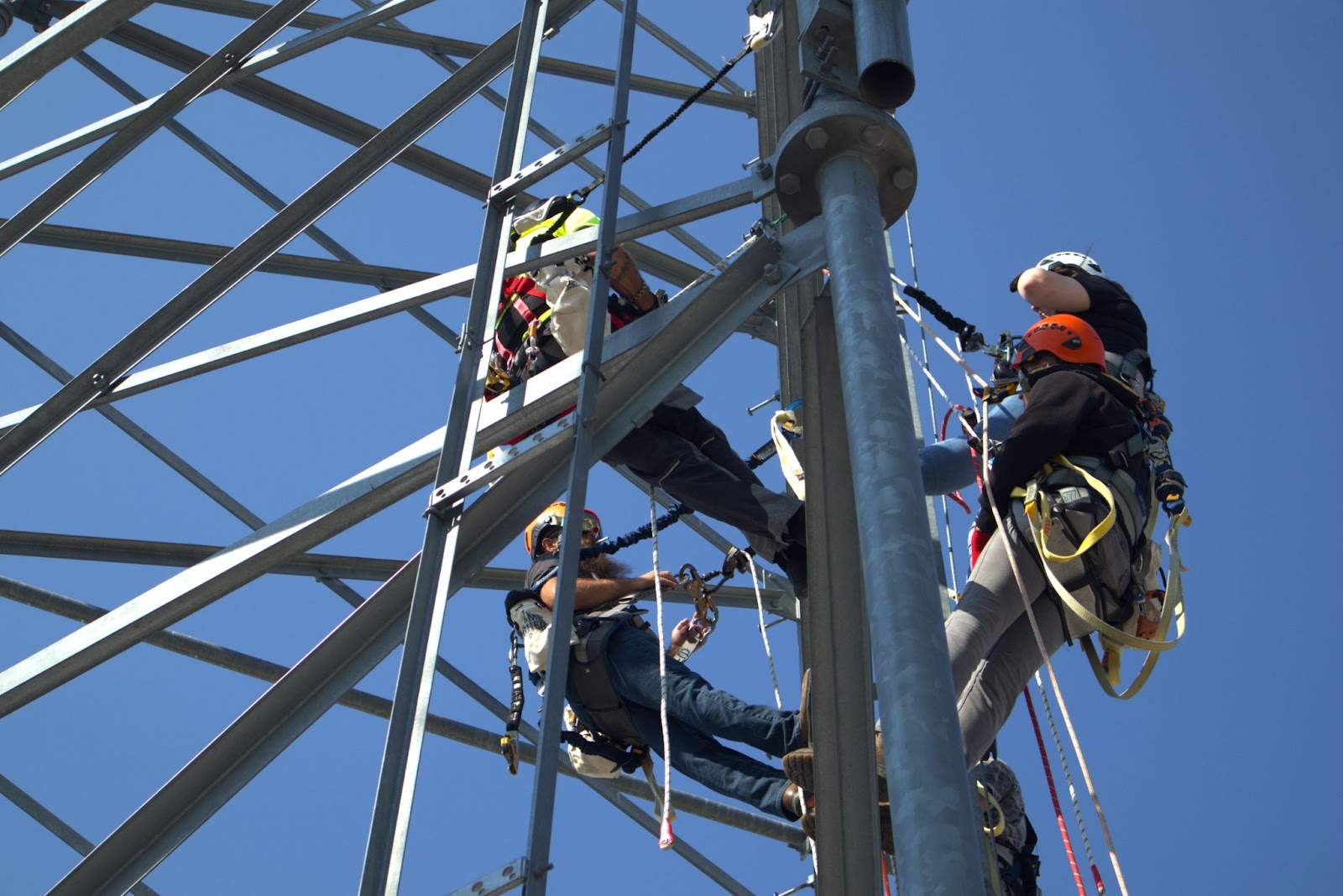 Tower Technician students practicing climbing, rigging, and safety techniques at the outdoor lab of Pinnacle Career Institute. The students are equipped with harnesses and safety gear, ascending a metal tower structure against a clear blue sky as part of their hands-on training.