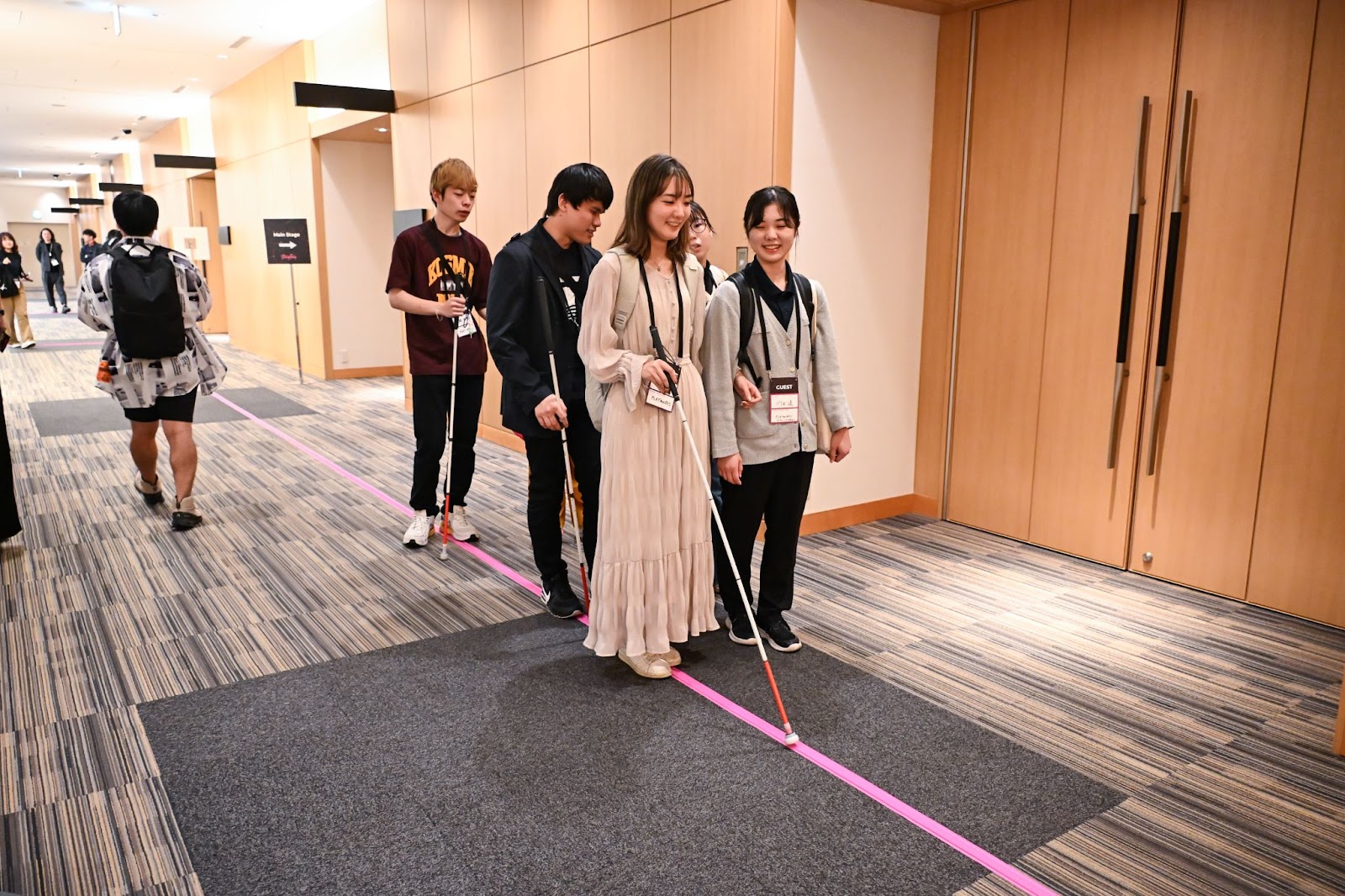 会場内の床に貼られたピンク色のココテープをガイドに歩く視覚障害のある学生たち。みんな笑顔で談笑している。