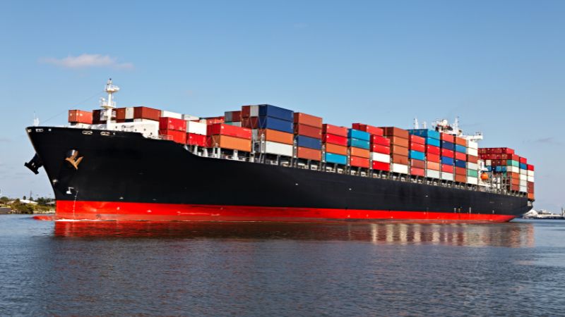 A large black-and-red container ship, loaded with multicolored containers, sails on calm waters under a clear blue sky.