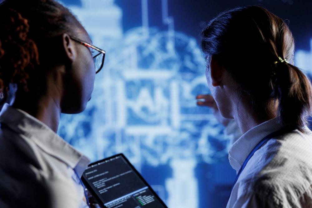 Two professionals analyzing data on a blue digital screen with graphical representations, while one holds a tablet displaying visible code.