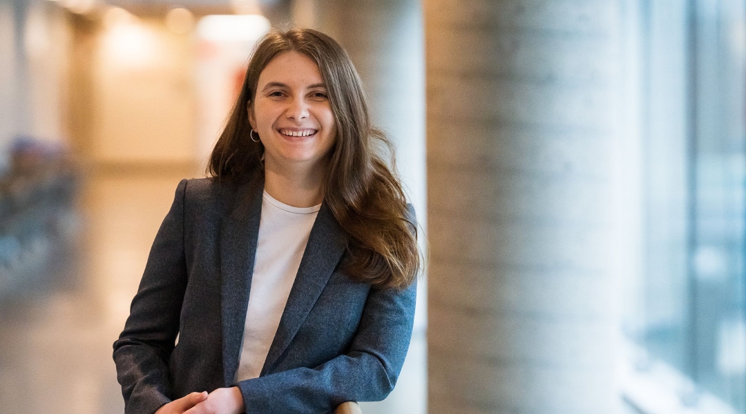 Smiling dietitian with long brown hair, wearing a blazer and white shirt, standing in a bright hallway with large windows.