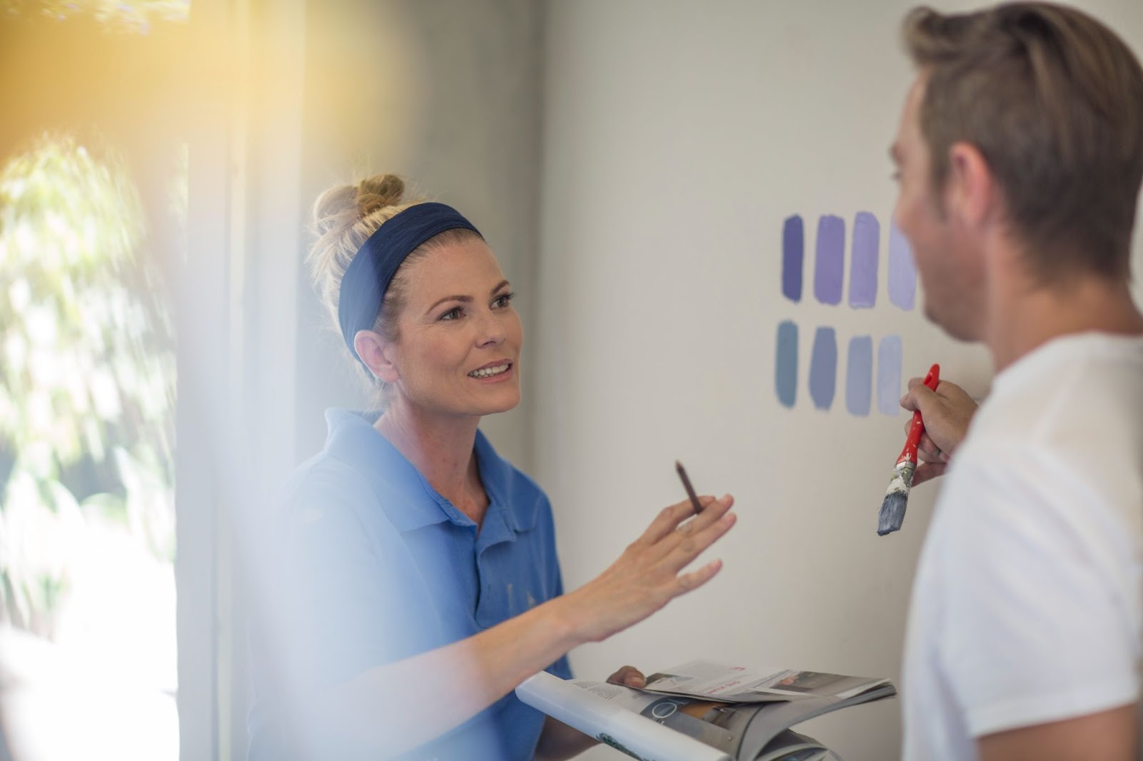 In a newly renovated room, a female client holding a brochure discusses paint color options on the wall with a male painter.