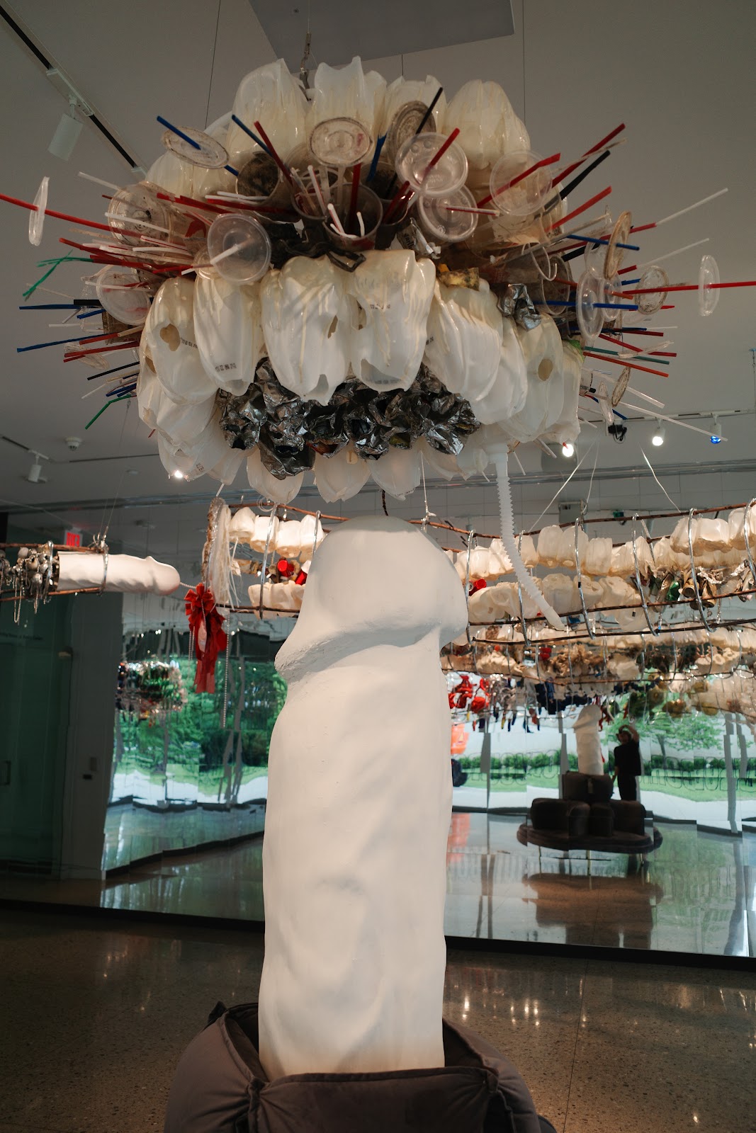 Plaster phallus sculpture pointing upwards towards the chandelier structure made of wire and plastic cups.
