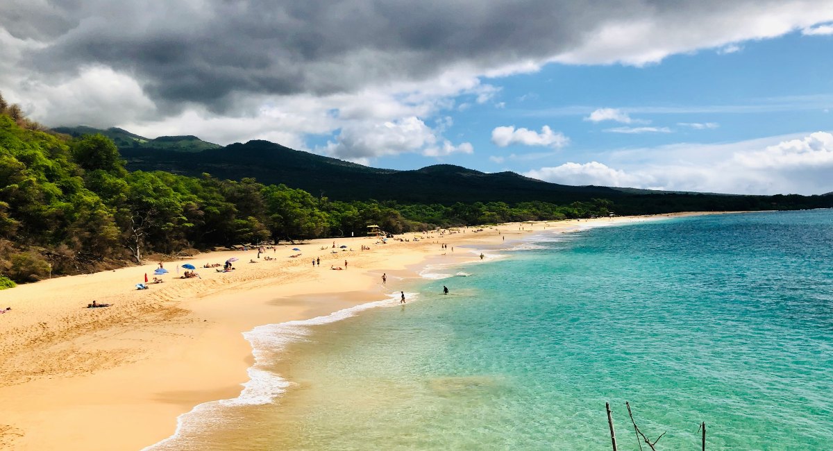 The Wailea-Makena area is home to one of the best Maui beaches, showcased here by a broad sandy beach with clear, shallow waters, framed by lush mountains under a dramatic cloudy sky.