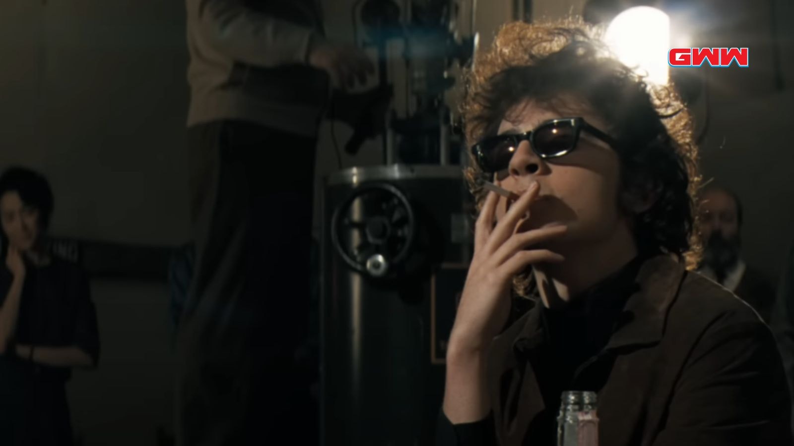 Bob Dylan with curly hair smokes while wearing sunglasses indoors.