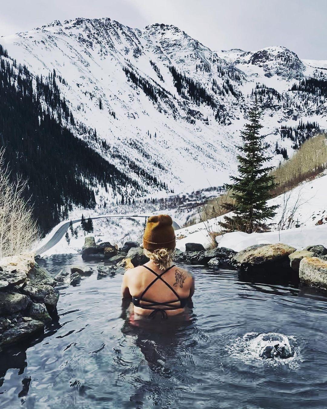 Woman in a black bikini with a brown hat in a natural pool in front of a snowy mountain landscape