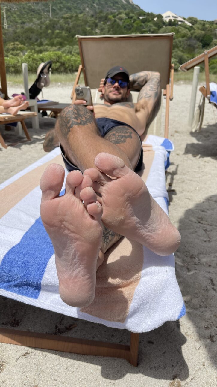 Papi Kocic posing shirtless at the beach on a beach towel taking photos of his bare feet for gay xxx onlyfans foot fetish content