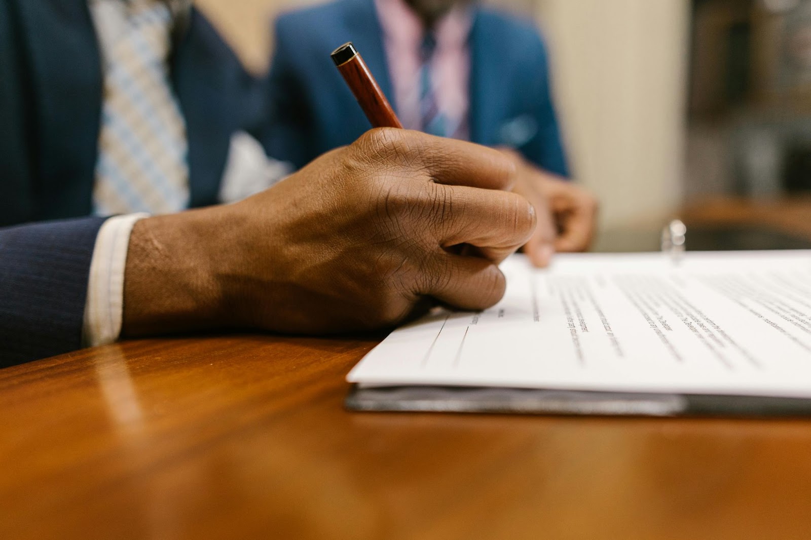 A person in a suit signing a document with a pen, with another person visible in the background.