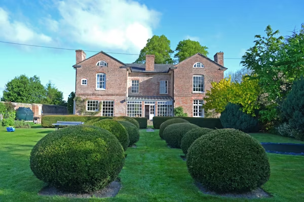 Historic brick estate with a formal garden featuring neatly manicured spherical shrubs, lush green lawn, and symmetrical landscaping design.