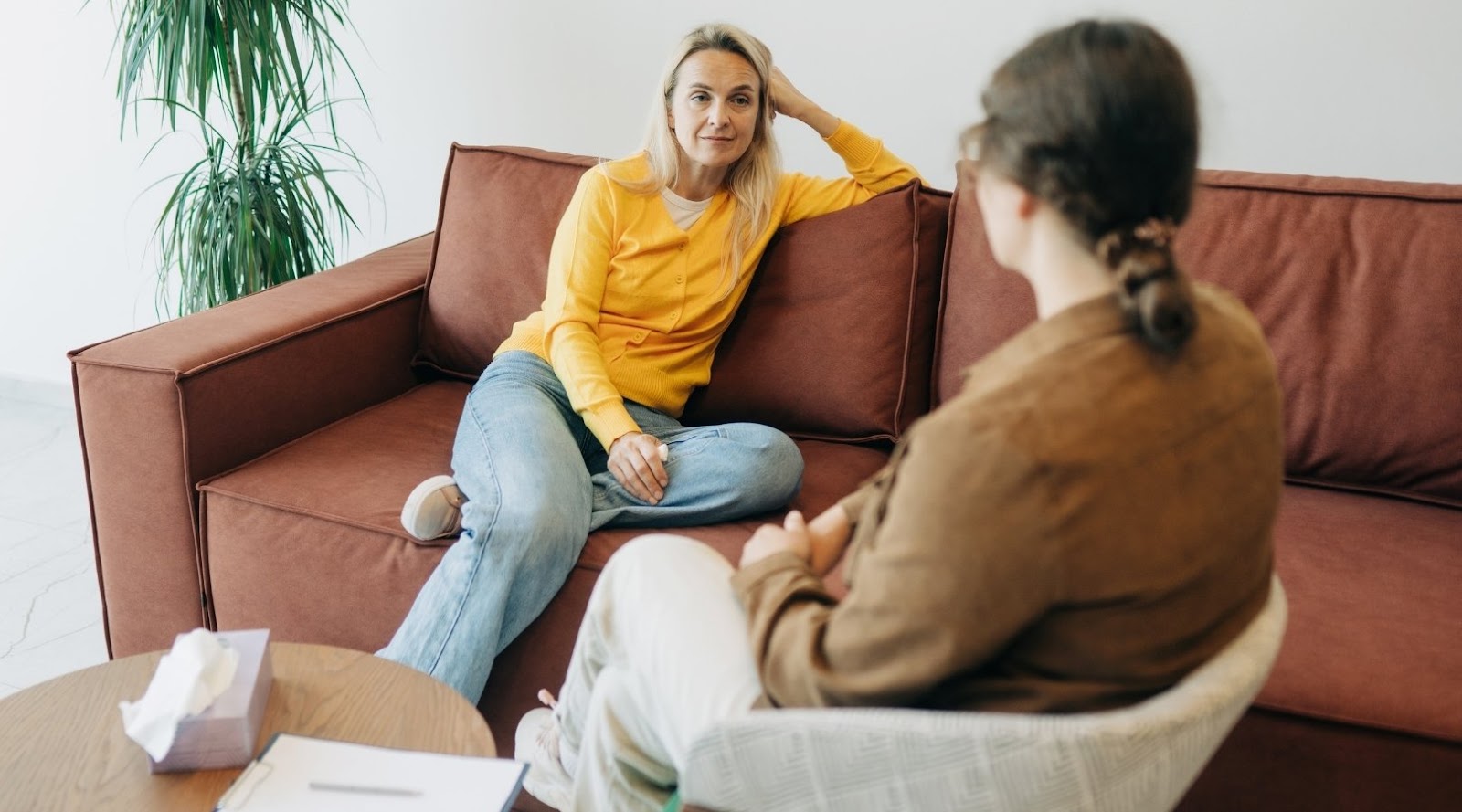 A dietitian and a client having a conversation, with one sitting on a couch wearing a yellow sweater, and the other sitting with her back to the viewer.