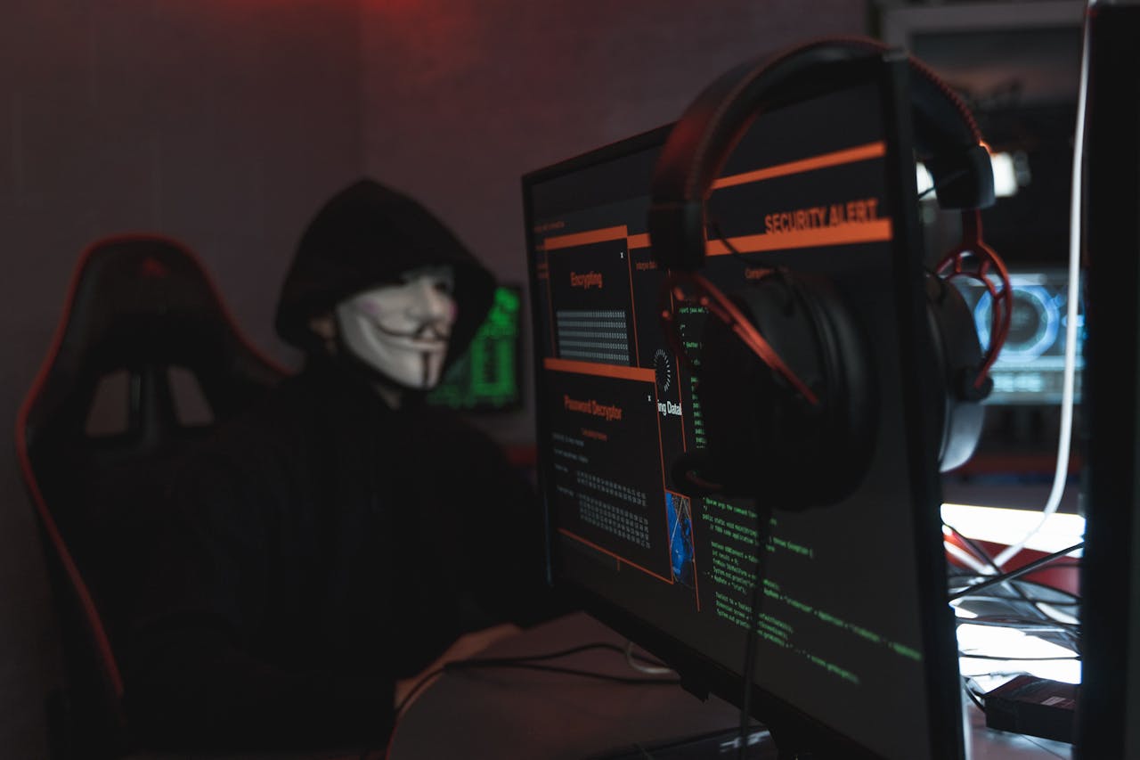 A person wearing a mask and hoodie sits in front of multiple computer screens displaying security alerts, symbolizing cybersecurity threats and the need to protect computer systems.