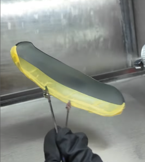 A gloved hand holding a 3D-printed transparent resin model with tweezers, applying a clear coat in a spray booth to enhance clarity and achieve a glass-like finish.