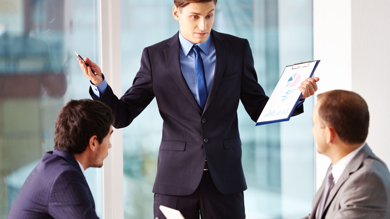 A businessman in a suit holding a clipboard and pen, presenting a report to two other individuals seated at a table during a business meeting.