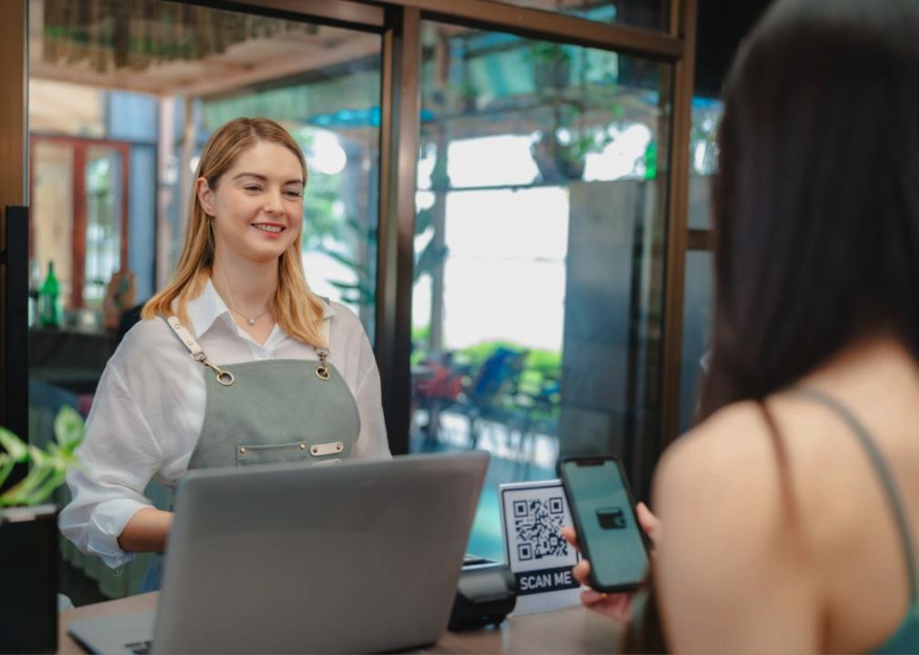 A smiling female cashier, wearing a light green apron is assisting a customer. The customer, seen from behind, is holding a smartphone and scanning a QR code displayed on the counter for payment. 