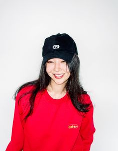 This contains a picture of Min Hee in a red shirt and black hat smiles at the camera while standing against a white wall