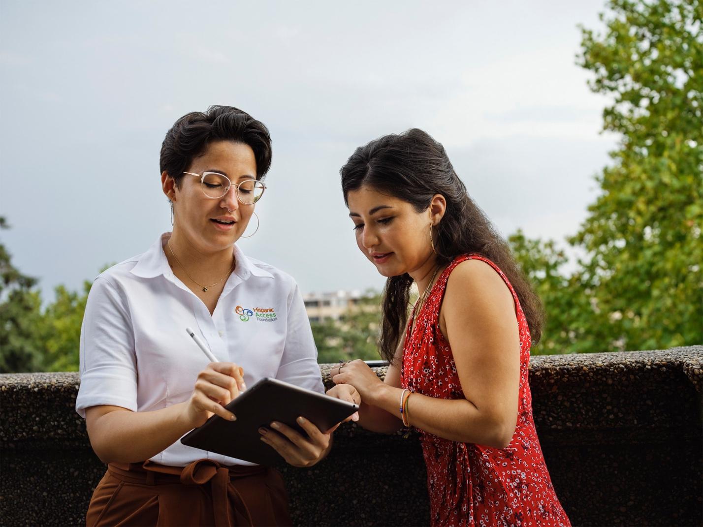 A person looking at a tablet with a pen and a person standing next to her

Description automatically generated