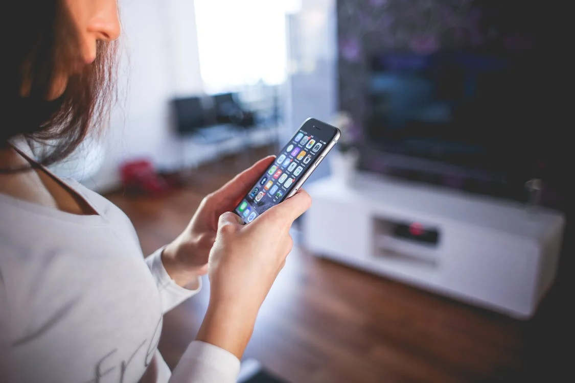 A person holding a smartphone with various app icons displayed on the screen, standing in a modern living room with a TV and furniture in the background