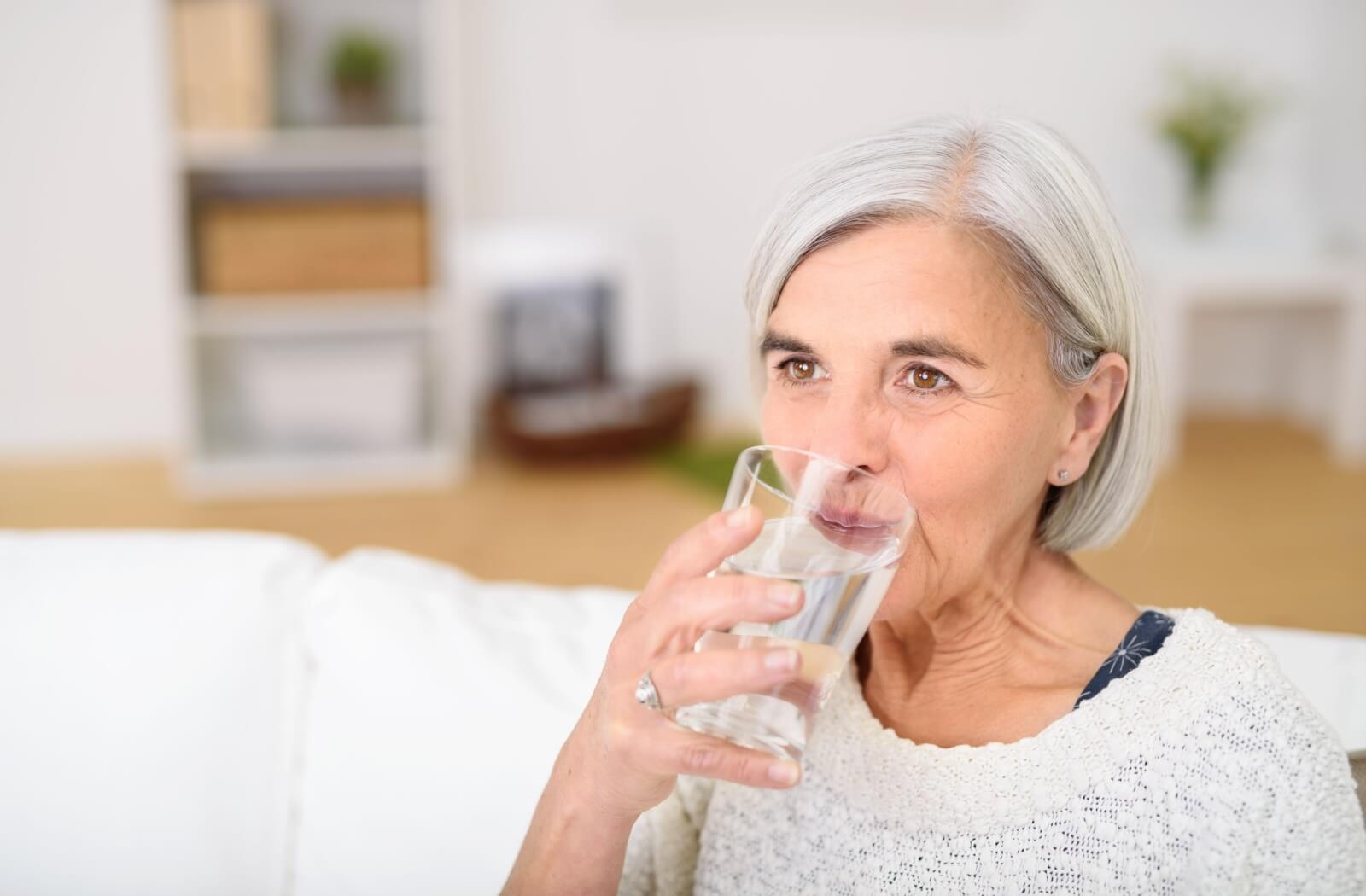 Older adult sitting on a couch and sipping a glass of water in a bright and comfortable living space