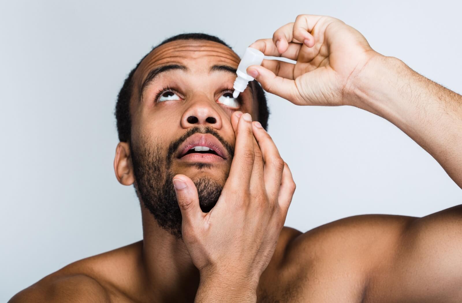 A bearded adult against a grey background holds their eyelid down to apply eye drops to relieve dry eye symptoms