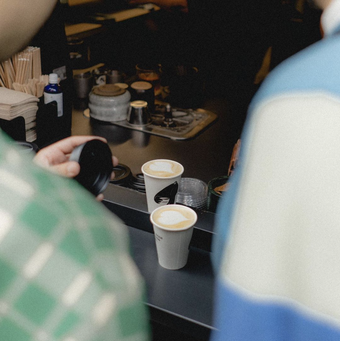 Two customers holding coffee cups with latte art at a café counter, with baristas preparing drinks in the background.