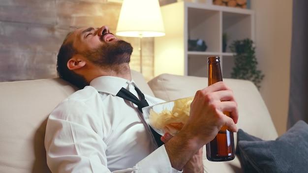 Businessman in formal wear eating chips from a bowl. Entrepreneur laughing watching tv.