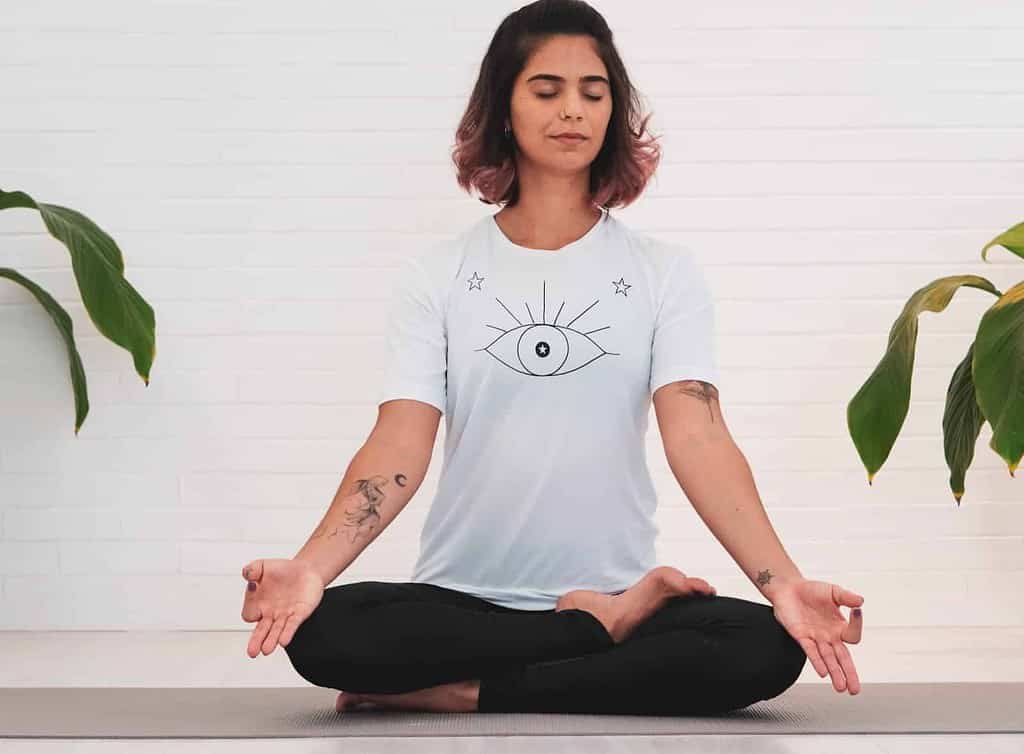 An individual sitting cross-legged on the floor in a meditative pose with hands resting on knees, palms facing upwards. The individual is wearing a white t-shirt with a graphic of an eye surrounded by star-like symbols and rays, paired with black pants. In the background, there is a plain wall and two potted plants on either side of the person.