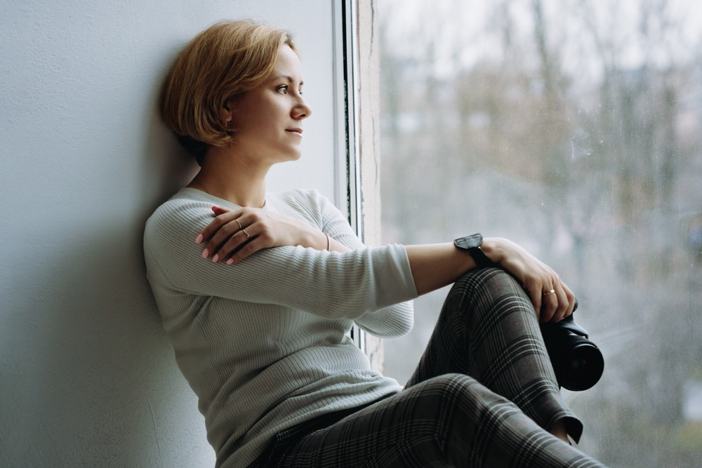 A woman is seen sitting beside a window, observing the outside view.