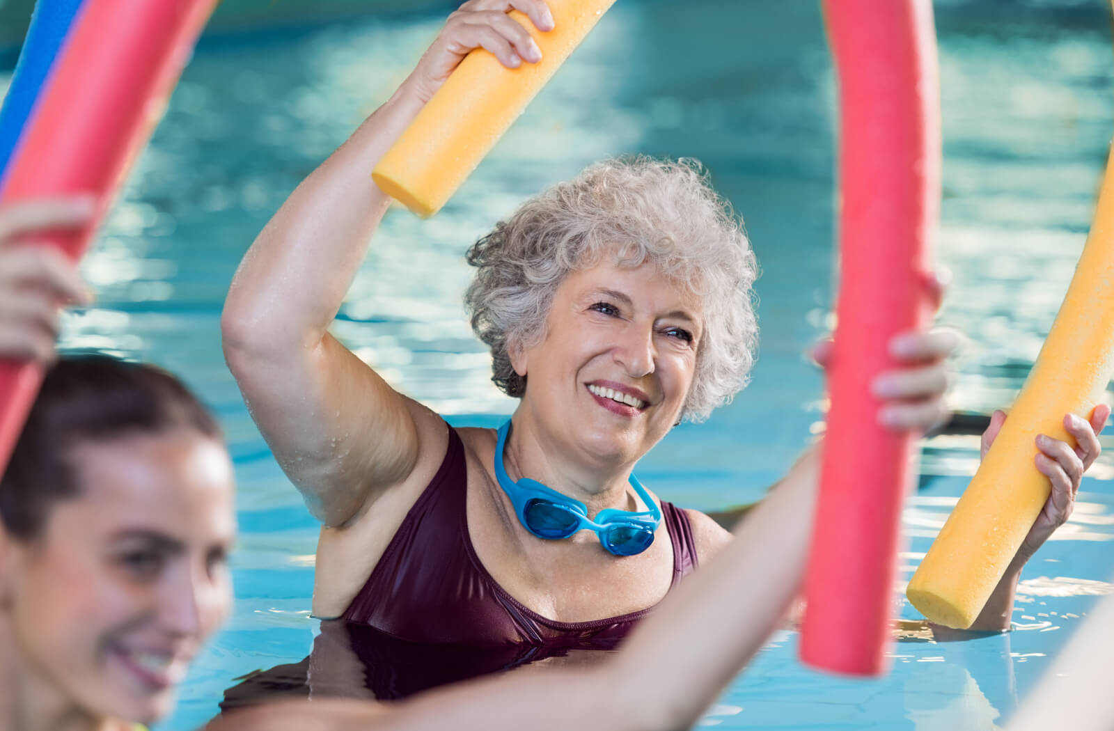 A smiling older adults holding a foam pool noodle overhead during a water aerobics class.