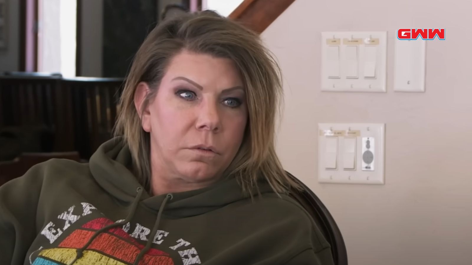 Meri Brown with short brown hair, wearing green hoodie, sitting near light switches.