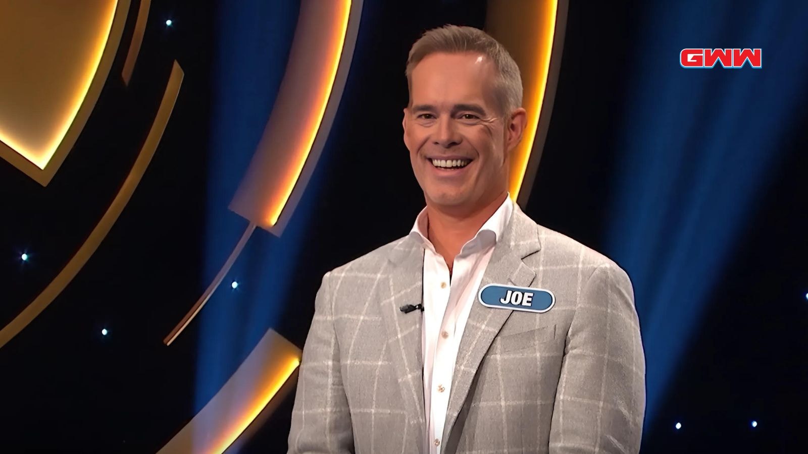 Contestant Joe smiling on a game show, wearing a light gray suit.