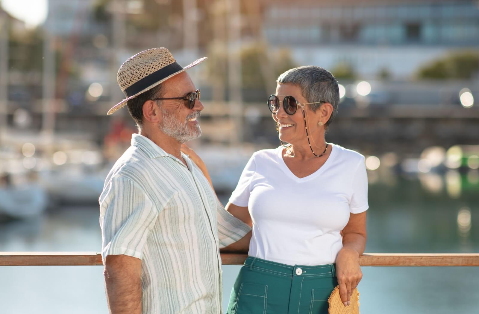 A senior couple smile and embrace each other while outside, wearing sunglasses to protect their vision.