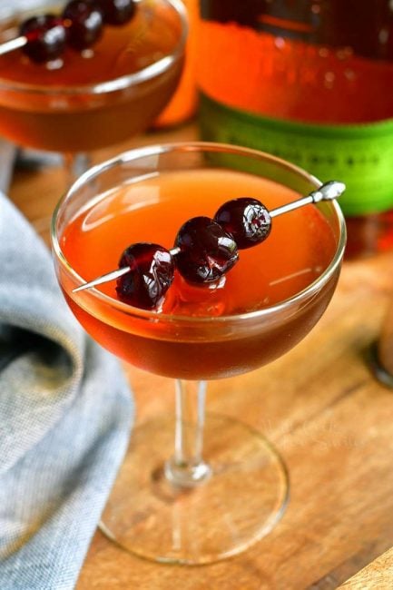 closer view of the light brown Manhattan cocktail in a glass topped with dark cocktail cherries on a silver pick