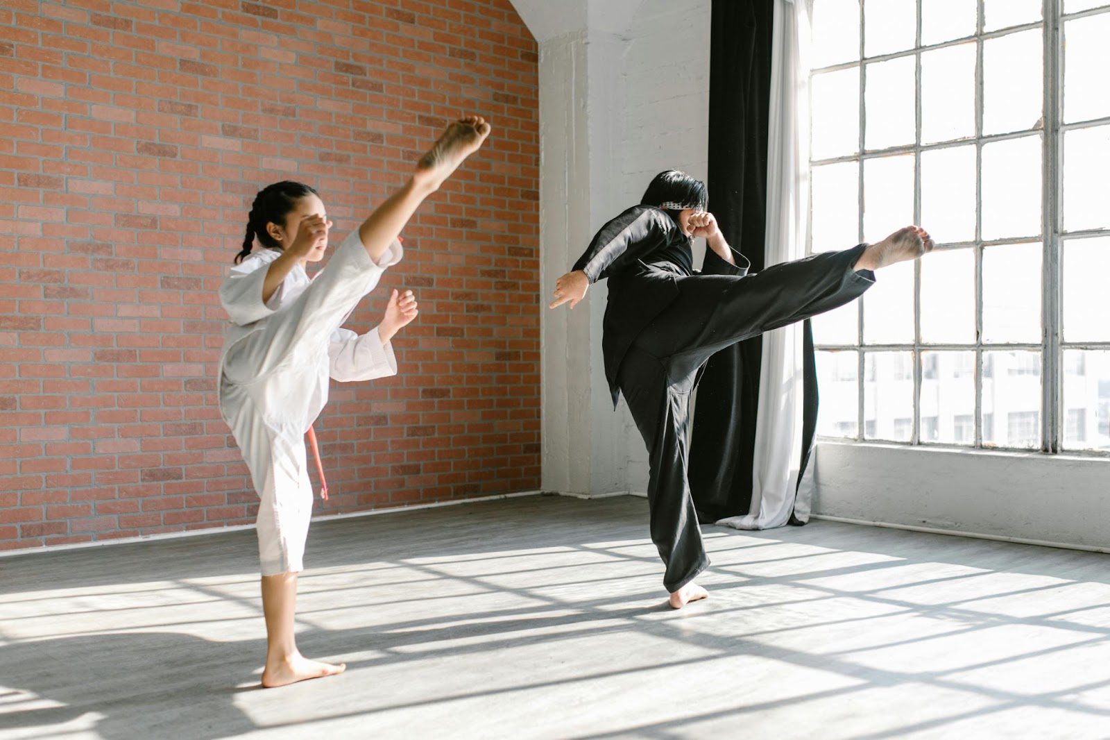 Two young martial arts students standing side by side, each mid-kick with one leg raised in the air.