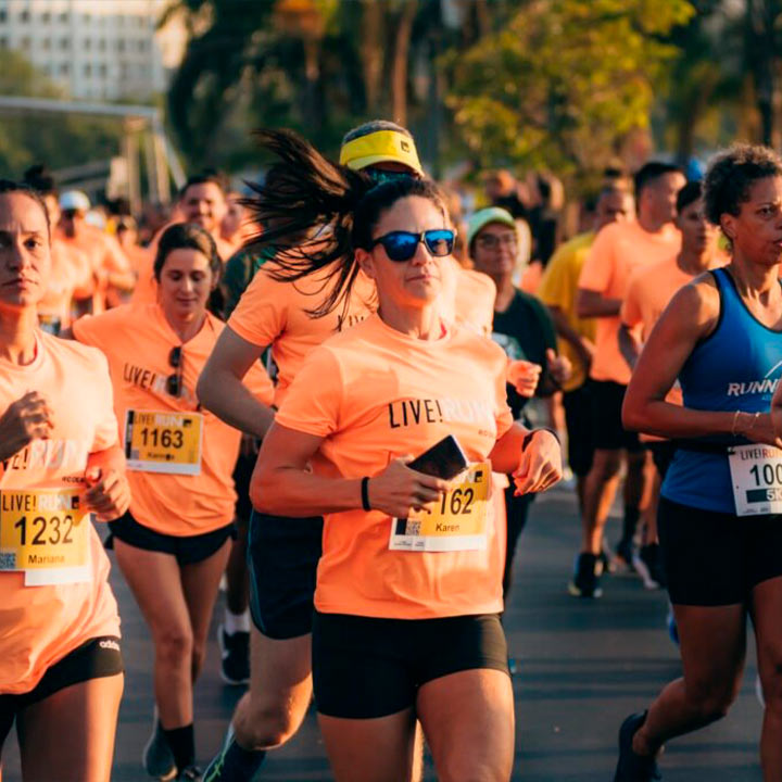 Grupo de corredores participando de uma maratona ao ar livre com roupas esportivas e expressões de esforço, numa manhã ensolarada.