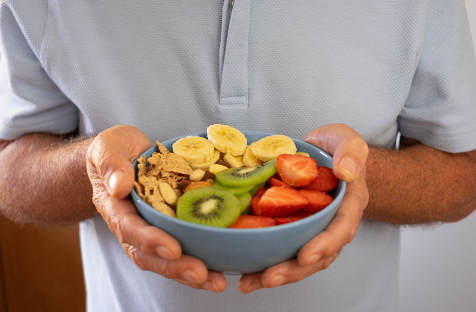 A senior holds an enticing bowl of kiwi, banana, and strawberry slices and cornflakes.