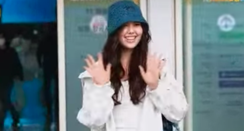 A picture of Olivia Marsh wearing a white jacket and a vest inside of it and a green cap and also waving her hand