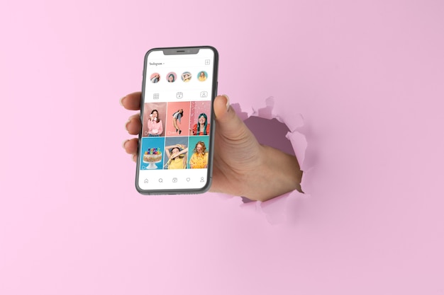 Hand breaking through a pink wall holding a smartphone displaying an Instagram profile with various posts