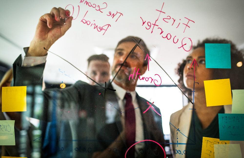 A team of professionals writing and analyzing graphs and notes on a transparent glass panel with colorful sticky notes attached.