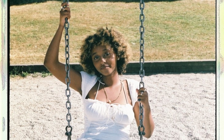 A Black femme with bleached hair sits on a swing, wearing a white dress