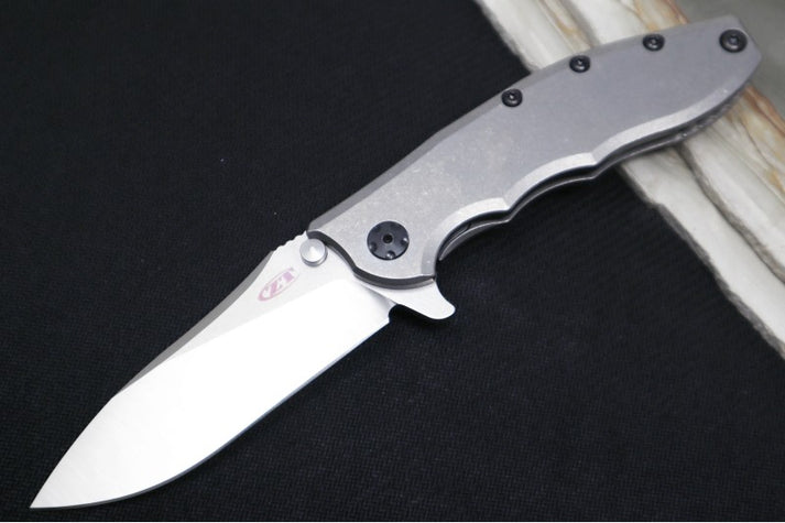 An image showcasing the Zero Tolerance 0562TI folding knife on a black and wood background. Designed by Rick Hinderer, it has a full titanium handle and a drop-point blade.