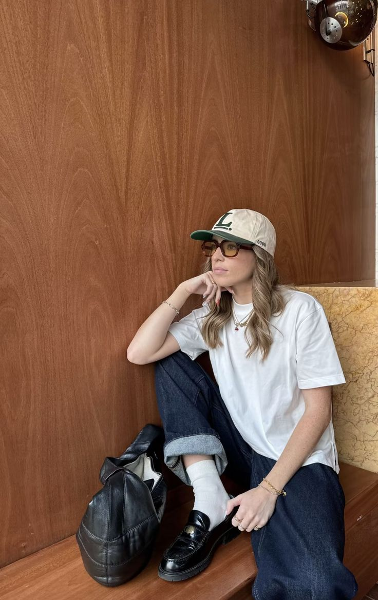 wanita duduk di kursi kayu memakai celana jeans biru, kaos putih, sepatu hitam, topi, dan kacamata