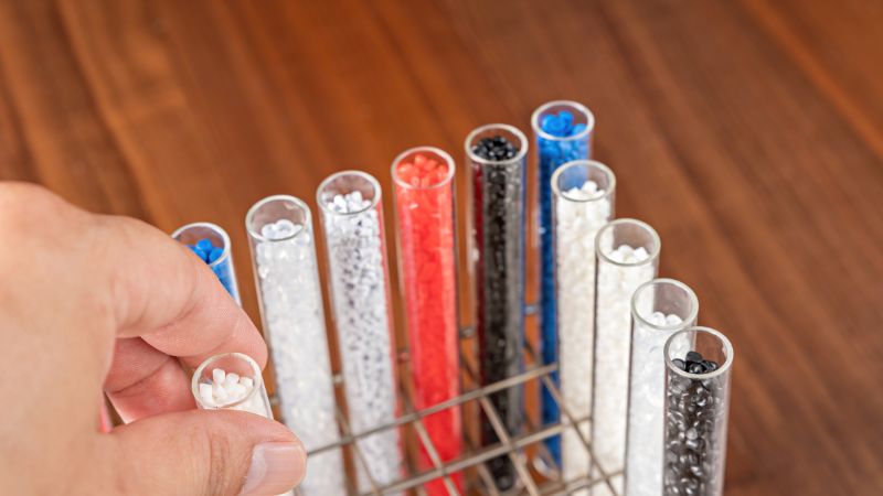 Selecting a test tube of small plastic pellets from a rack of colorful tubes on a wooden surface.