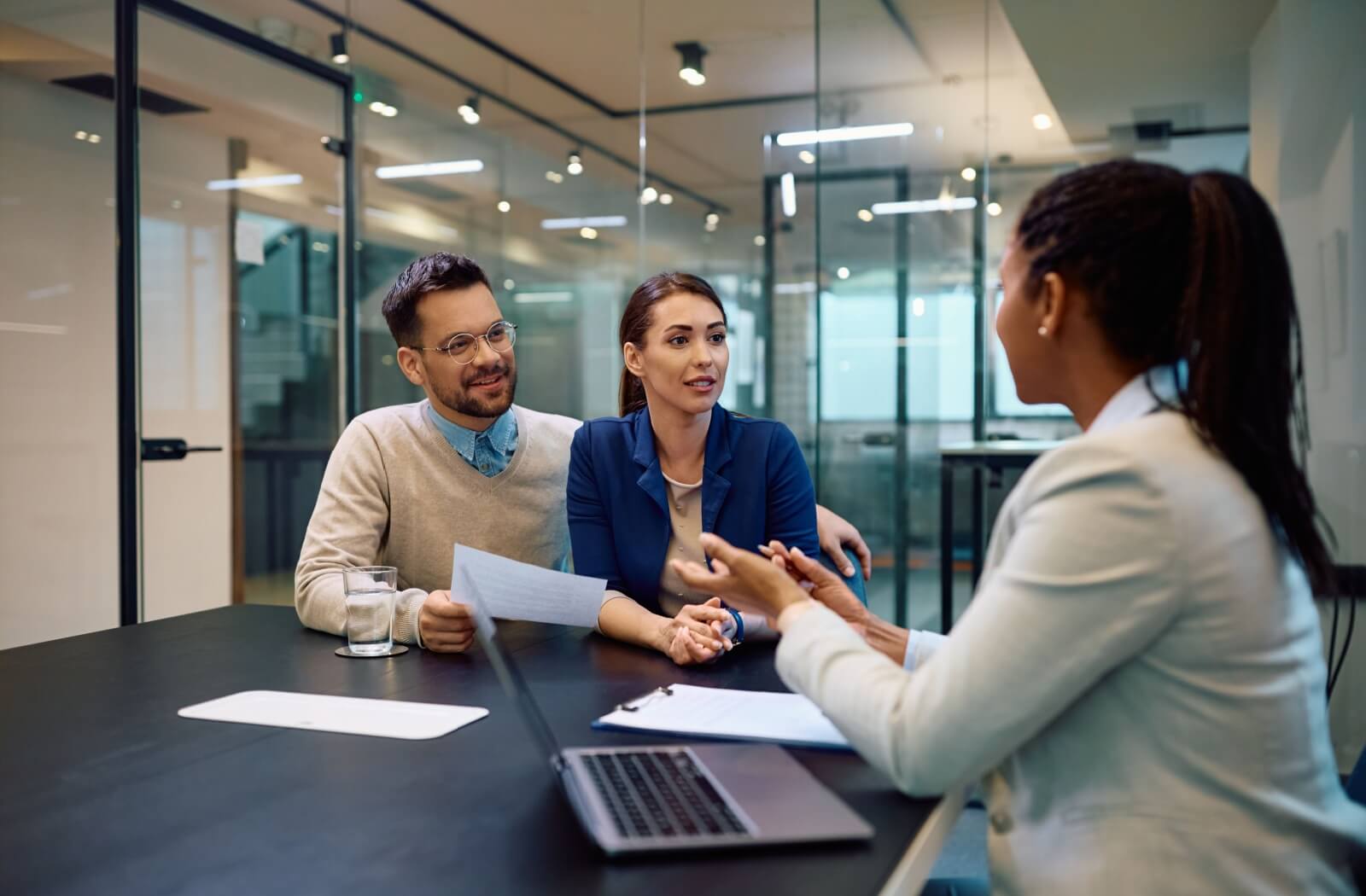 A couple sitting at a desk in a modern office, talking to a financial advisor with papers and a laptop on the desk in front of them