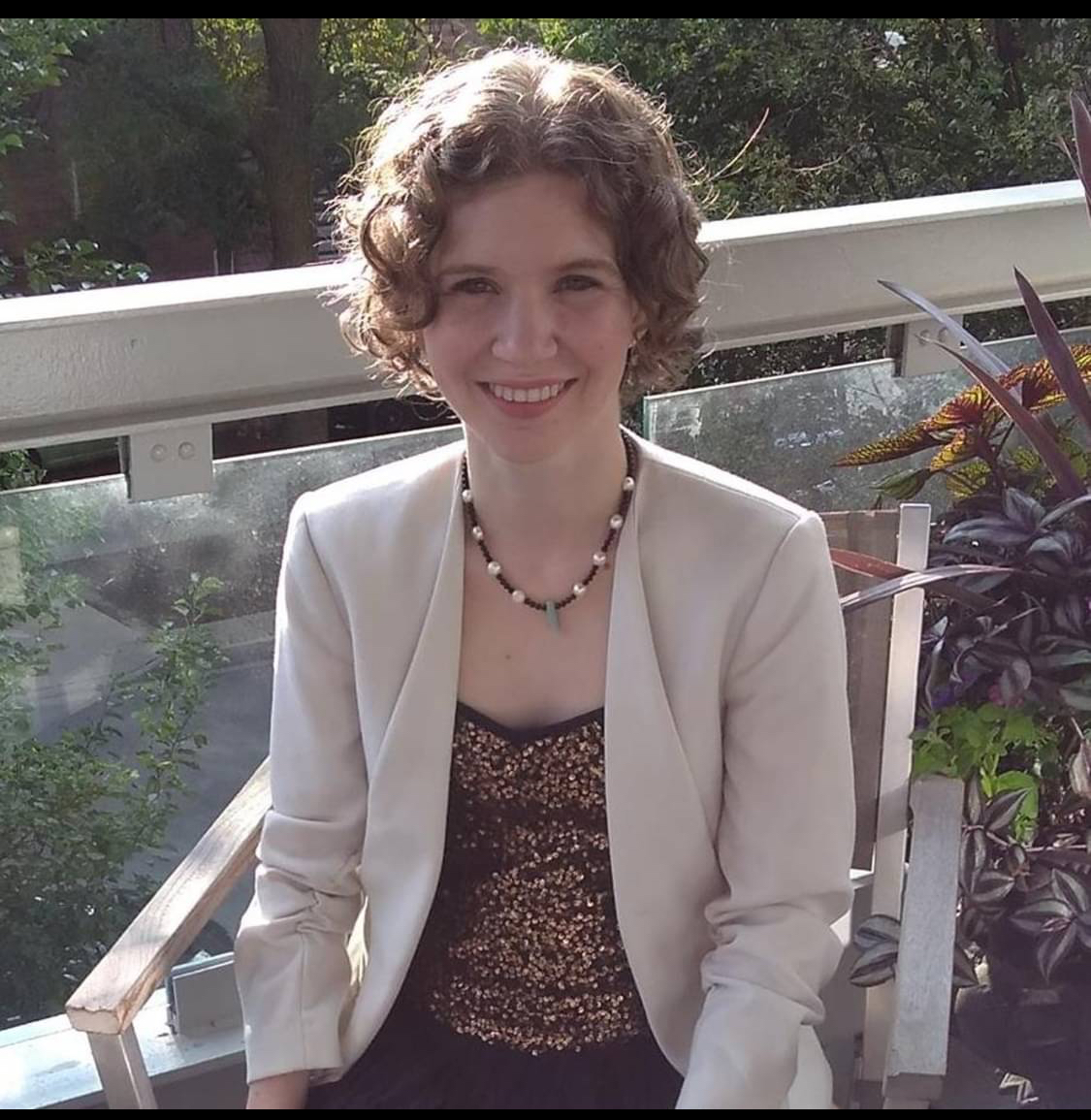 A woman poses in business attire. Greenery and a balcony wall in the background. 