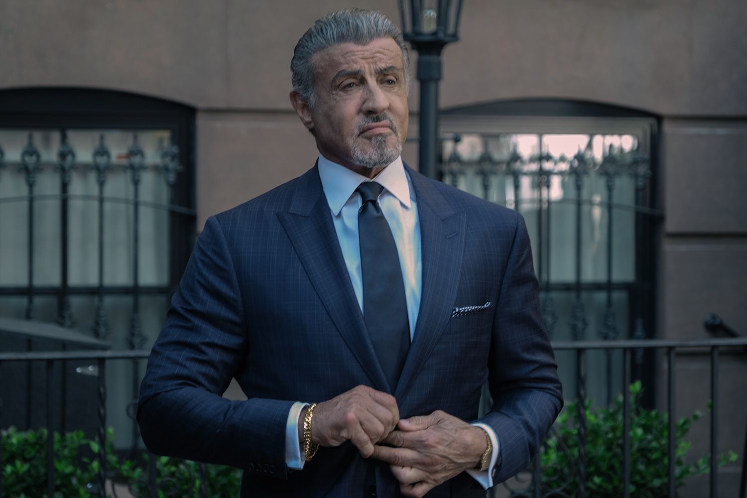 Sylvester Stallone con traje azul oscuro y corbata, posando con seriedad frente a un edificio elegante.