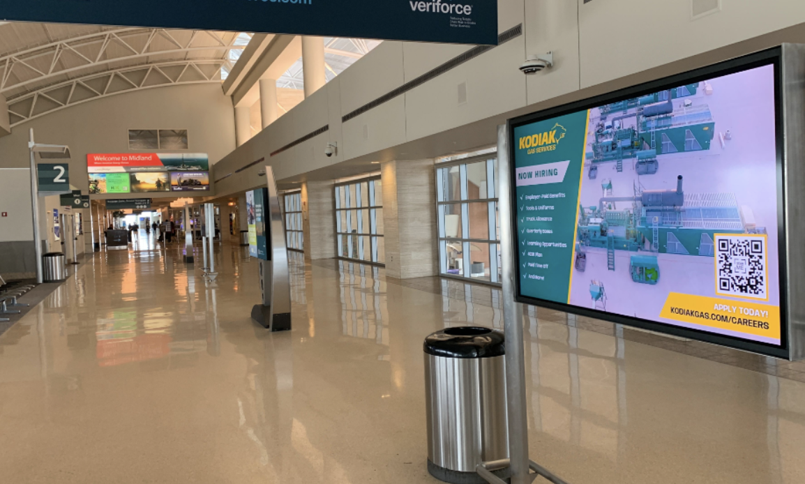 airport terminal hallway with a row of TV screen advertisement separating the walk space, as well as a hanging advertising banner in the distance.